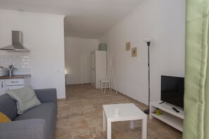 A modern living room with a grey sofa, a white coffee table, a flat-screen TV on a stand, a kitchen area with a stove and cabinets, and a hallway leading to another room.