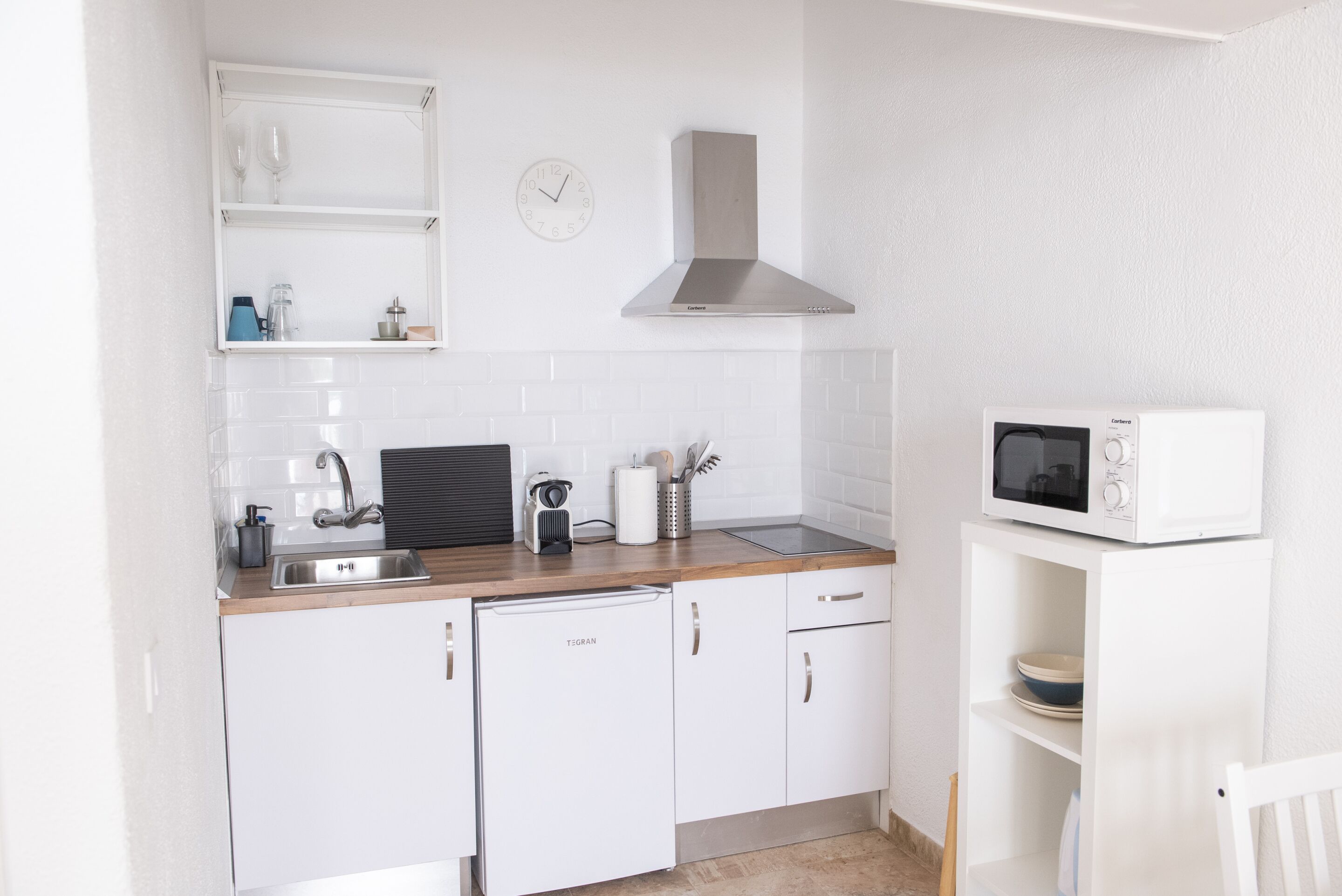 Une cuisine compacte avec des meubles blancs, une hotte en acier inoxydable, un four à micro-ondes et une machine à café.