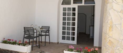 Une terrasse couverte avec une table et des chaises, un mur en pierre et une porte donnant sur un espace intérieur.