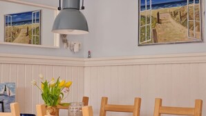 Un comedor con mesa de madera, sillas, una lámpara colgante y dos cuadros enmarcados con paisajes costeros.