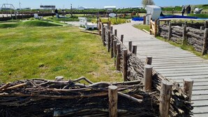 Una pasarela de madera que conduce a una zona de playa con un campo cubierto de césped y algunas construcciones al fondo.