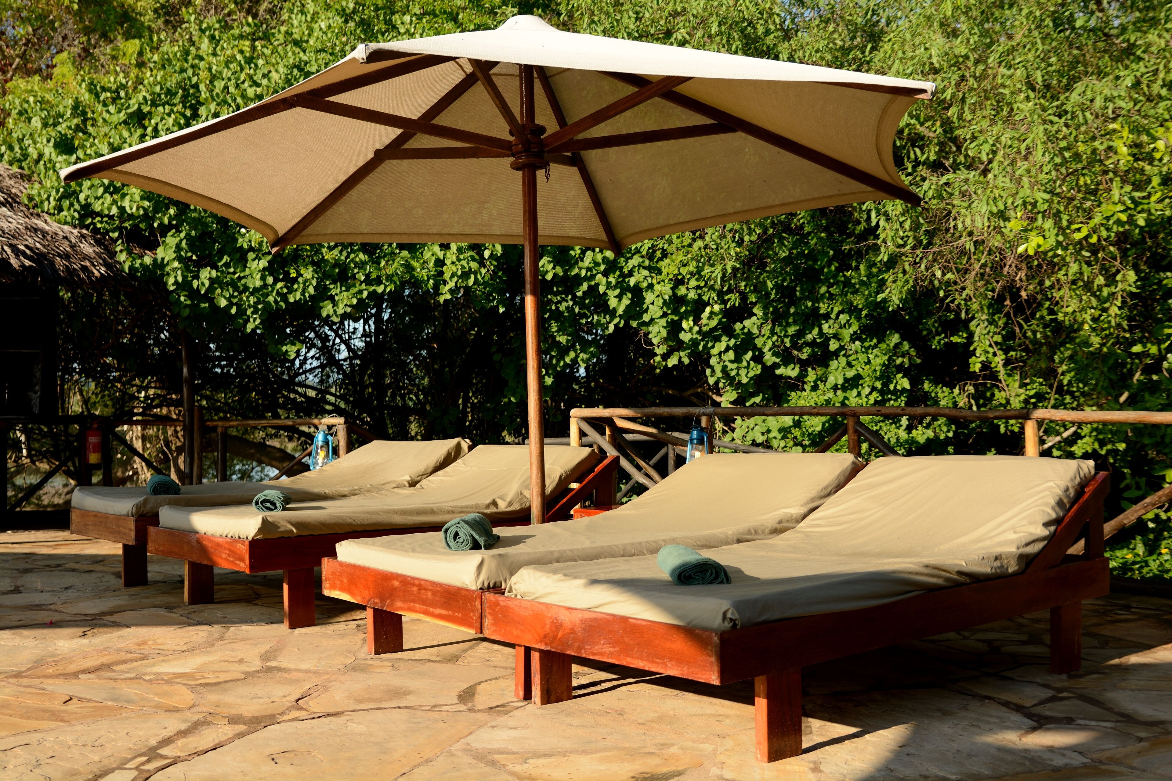 Un set di poltrone in legno con cuscini beige, disposte sotto un ombrellone grande, su un patio in pietra con uno sfondo di vegetazione verdeggiante.