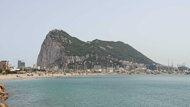 A coastal cityscape with a prominent rocky hill, a beach, and a marina with numerous boats.