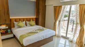 Habitación de hotel con una cama grande, cabecera de madera, mesita de noche con artículos de aseo y vistas a la ciudad a través de grandes ventanales.