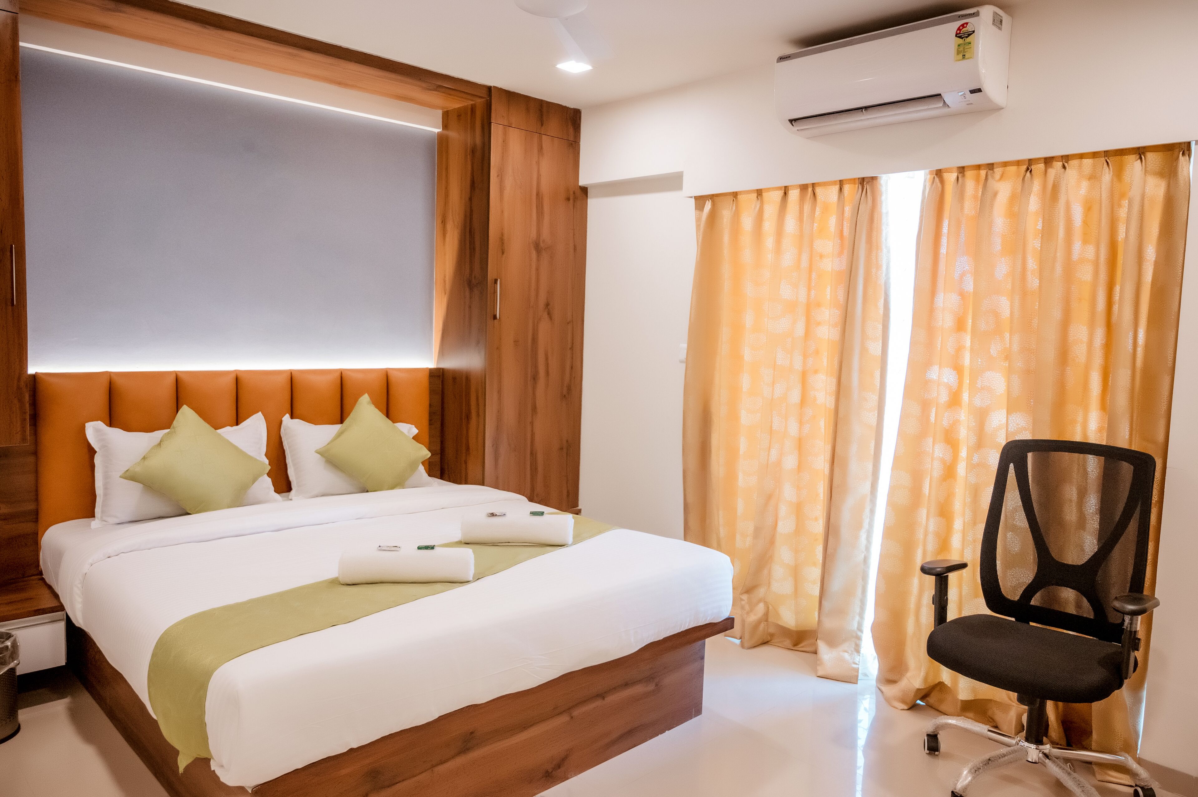 A hotel room with a wooden headboard, a bed with white linens, a desk with a chair, and a window with curtains.