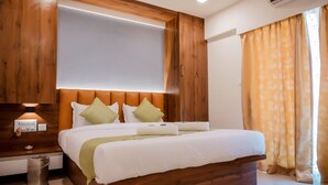 Habitación de hotel con cabecera de madera, cama con ropa blanca y almohadas verdes, mesita de noche con lámpara y ventana con cortinas amarillas.