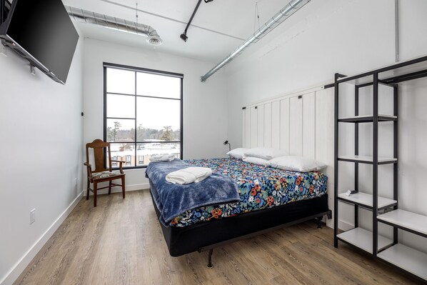 A modern bedroom with a large bed, a chair, a TV mounted on the wall, and a window with a view of trees.