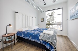 A bedroom with a bed, a nightstand, a radiator, a window with a view, and a painting on the wall.