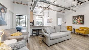 Una sala moderna con un sofá gris, sillones estampados y sillas amarillas. Hay una zona de cocina con un ventanal grande, y obras de arte en las paredes.