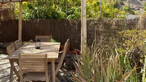 Een houten tafel en stoelen buiten, onder een rieten dak, omgeven door planten en met een bergachtige achtergrond.