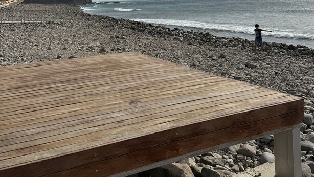 Een houten terras aan een rotsachtig strand, met een persoon in de verte.