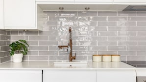 A kitchen with a white countertop, a sink with a brass faucet, and a backsplash made of gray tiles. There are three white containers on the counter. - Modern Apartment in Walthamstow (London)
