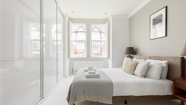 A bedroom with a large bed, a bedside table, a lamp, and a window with a view of houses. - Modern Apartment in Walthamstow (London)