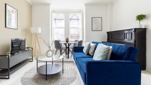 A modern living room with a blue sofa, a TV, and a dining area with a table and chairs. - Modern Apartment in Walthamstow (London)