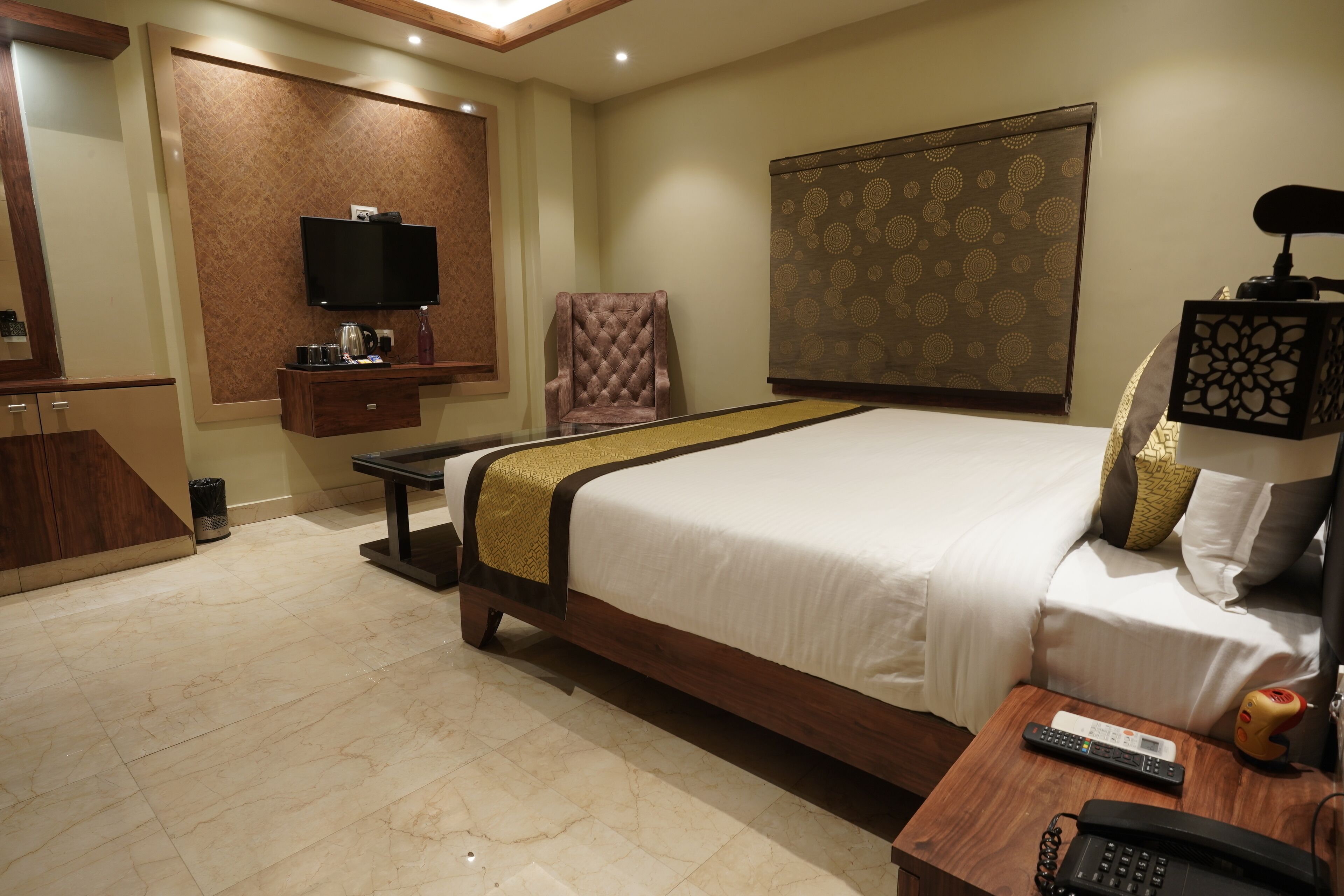A hotel room with a large bed, a flat-screen TV mounted on the wall, a wooden nightstand with a phone and remote, and a small table with a coffee maker and a bottle.