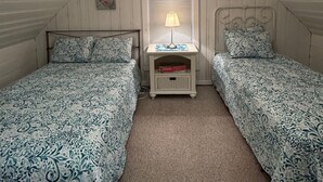 Un dormitorio con dos camas, un ventilador de techo, una mesita de noche con lámpara y una ventana con persianas.