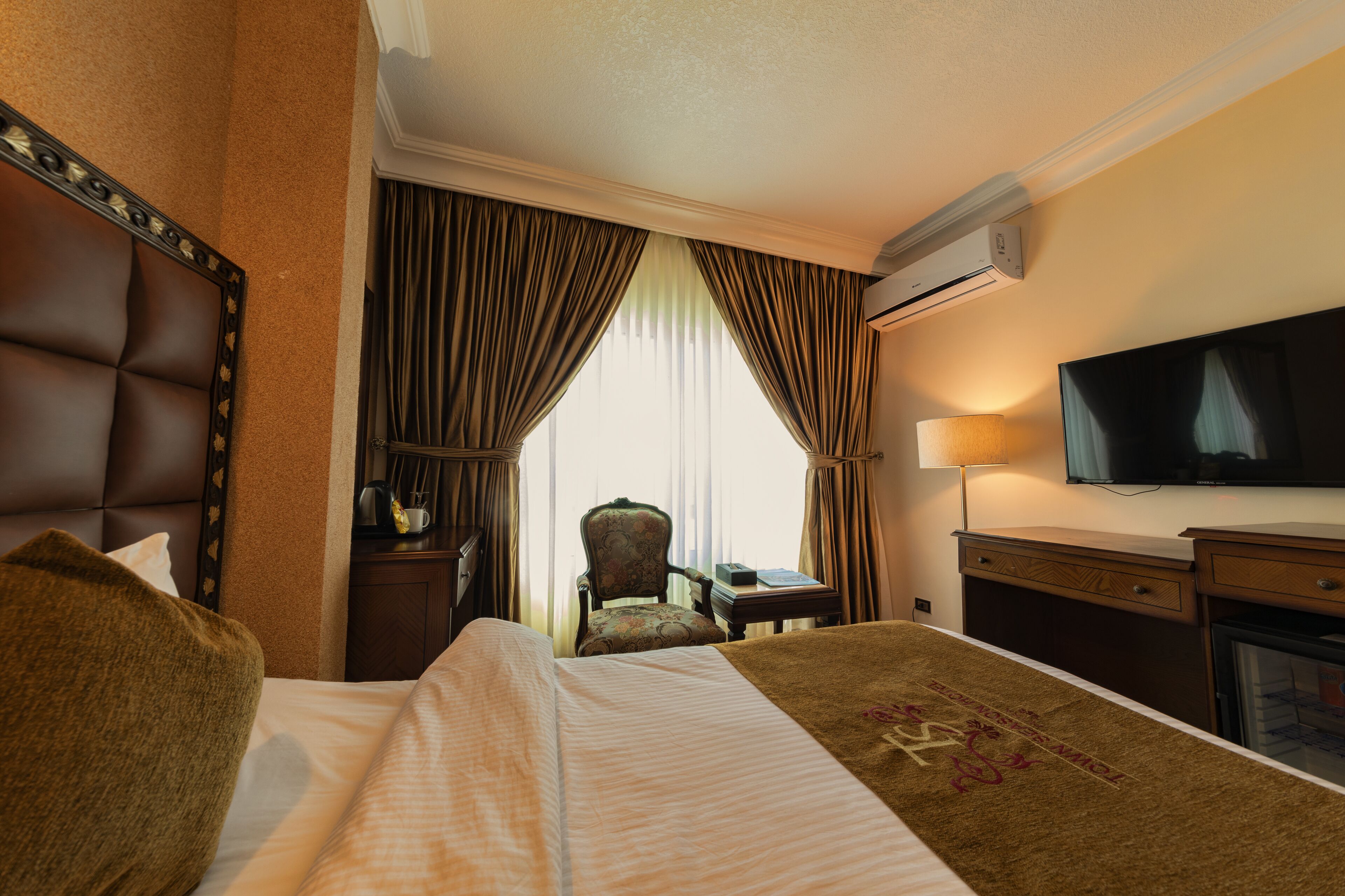 Quarto de hotel com cama, poltrona, escrivaninha, aparelho de ar condicionado e televisão.