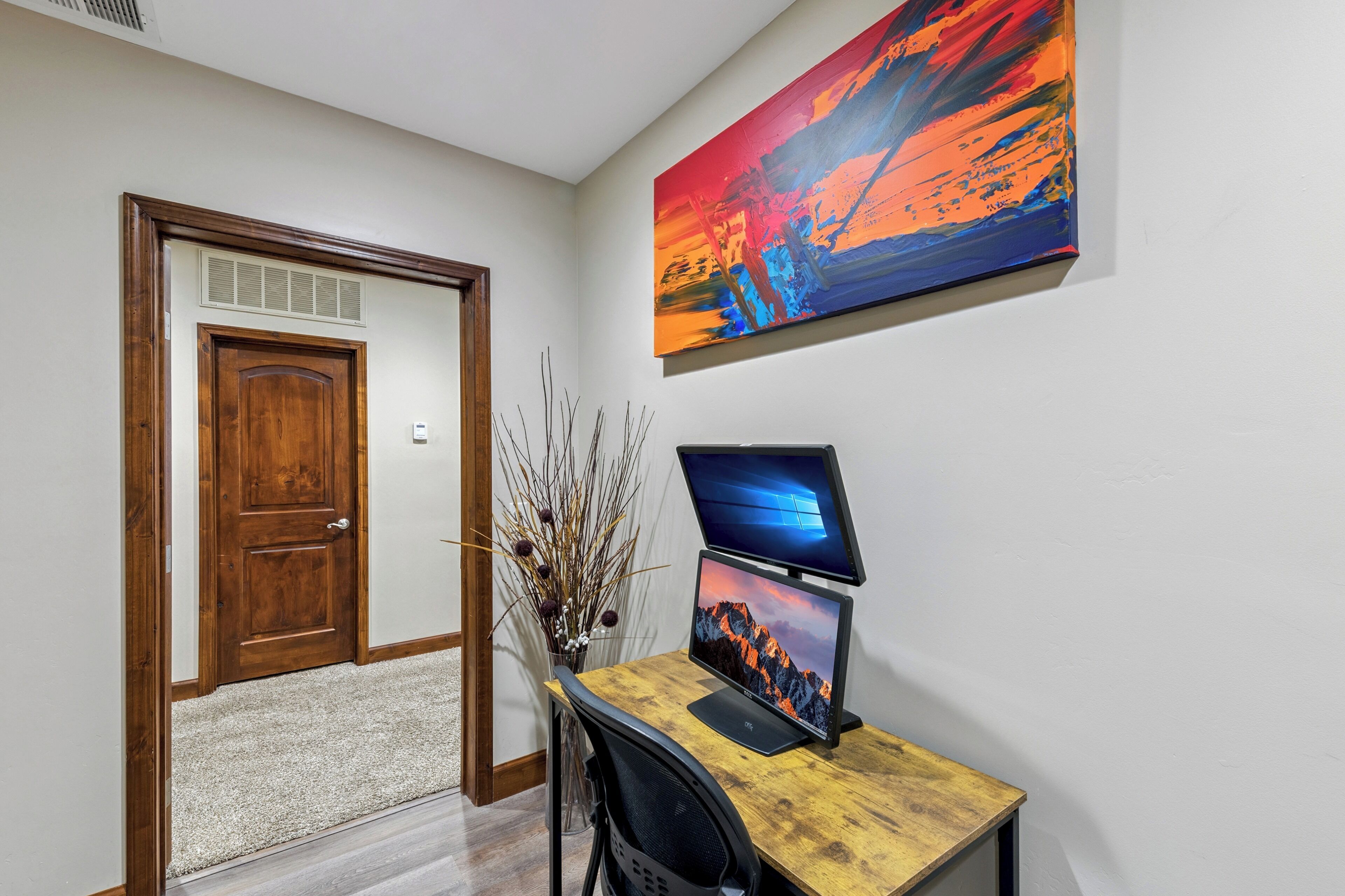 Una habitación con un escritorio de madera, una computadora, una puerta y un cuadro en la pared.