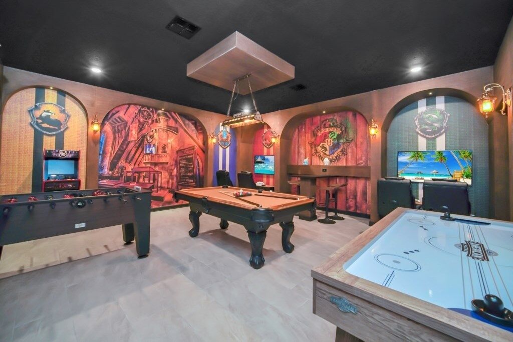 Mesa de bilhar, mesa de pebolim e área de bar com jogos de fliperama num espaço com tetos abobadados e arte decorativa temática.