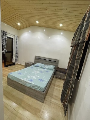 A bedroom with a wooden ceiling, a bed with a floral bedspread, a nightstand, and a window with curtains. - Résidence BANA (Ngaparou)