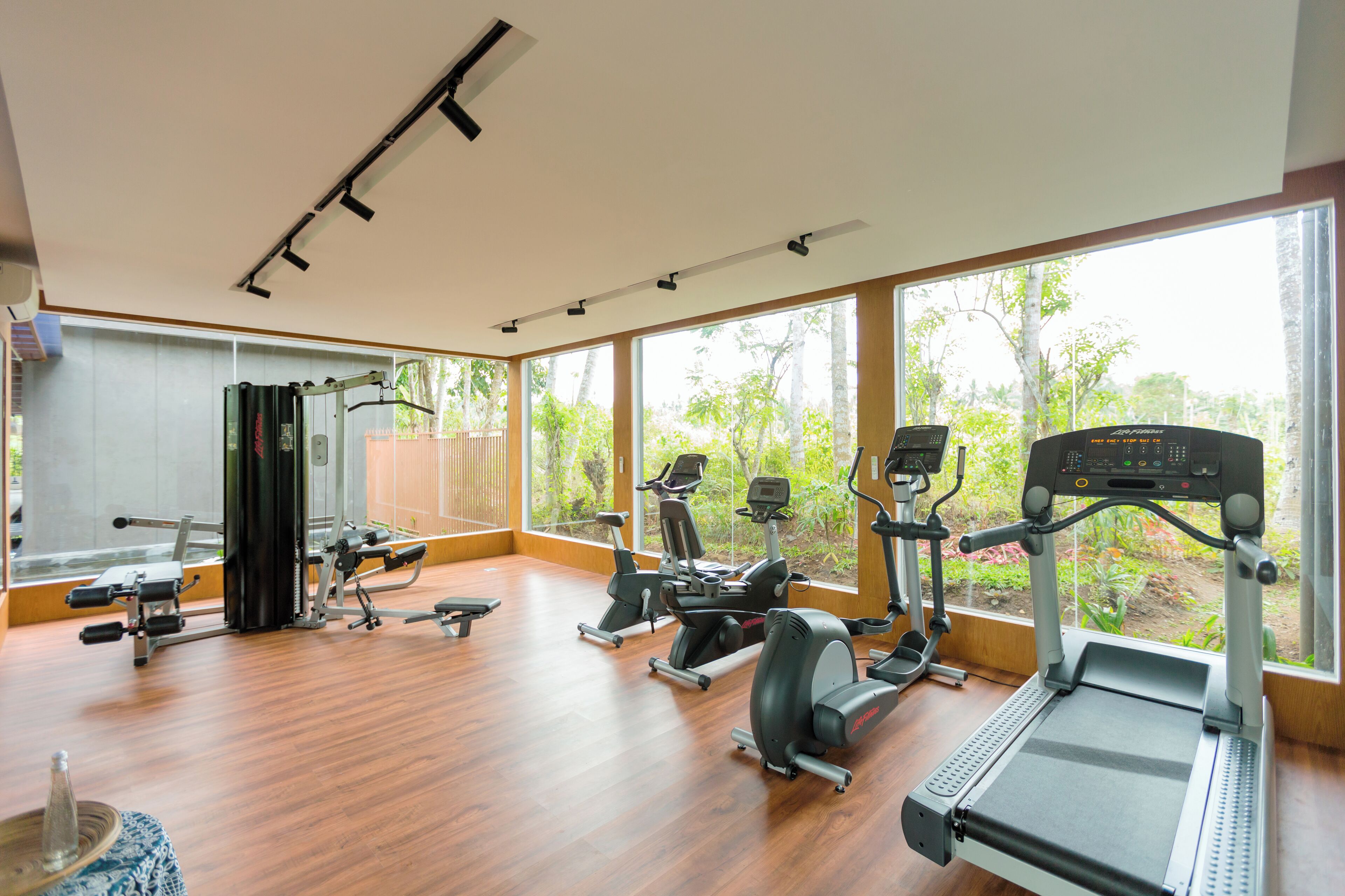 A spacious gym with various exercise equipment, large windows, and a wooden floor.