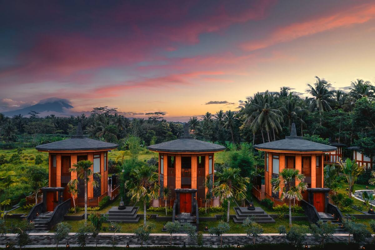 Un resort con tres edificios circulares, rodeado de exuberante follaje y palmeras, bajo un cielo colorido.