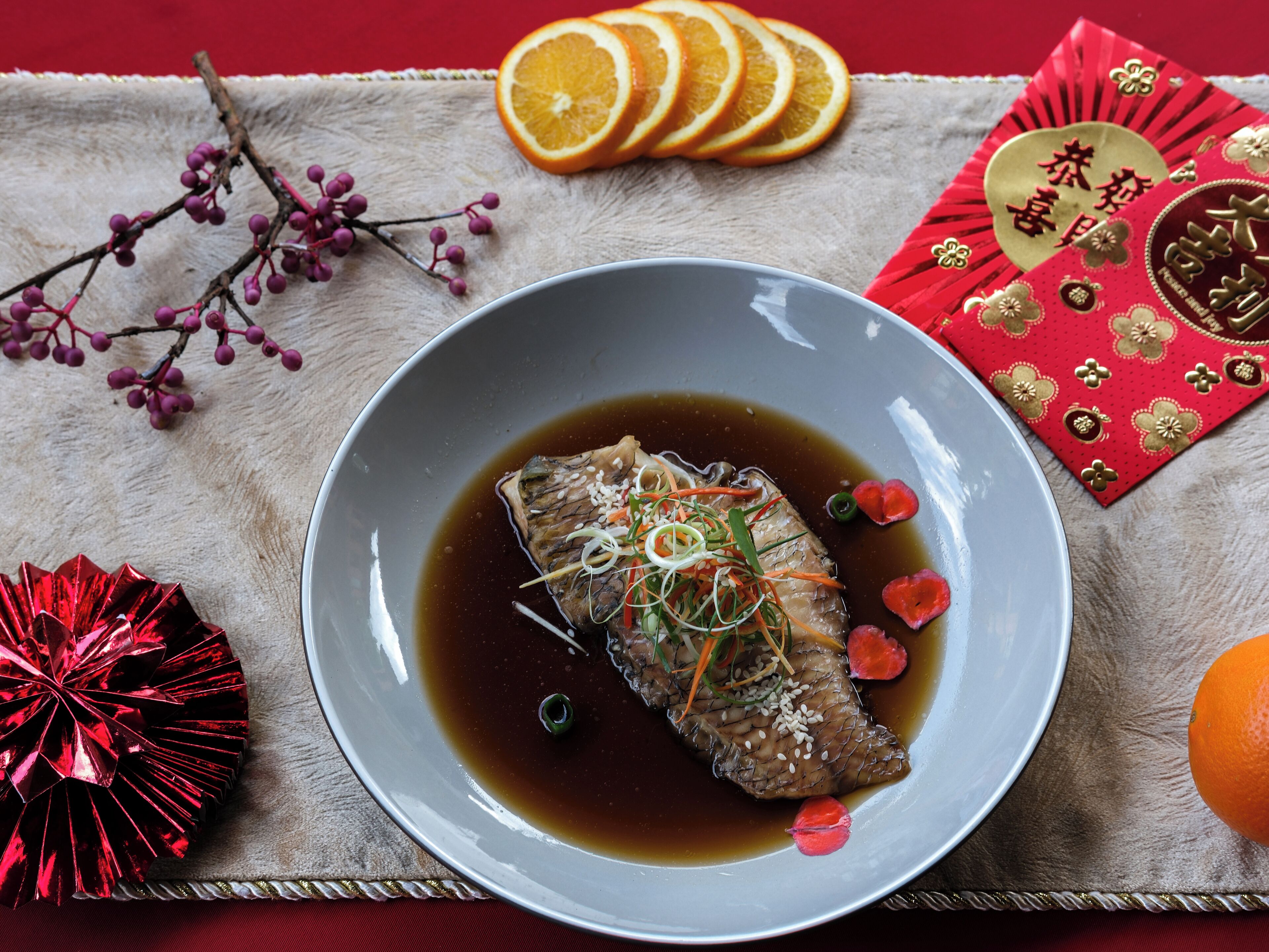 Un plat de poisson accompagné de légumes et de sauce, agrémenté d’éléments décoratifs tels que des tranches d’orange séchées, un éventail rouge et une branche ornée de baies violettes.