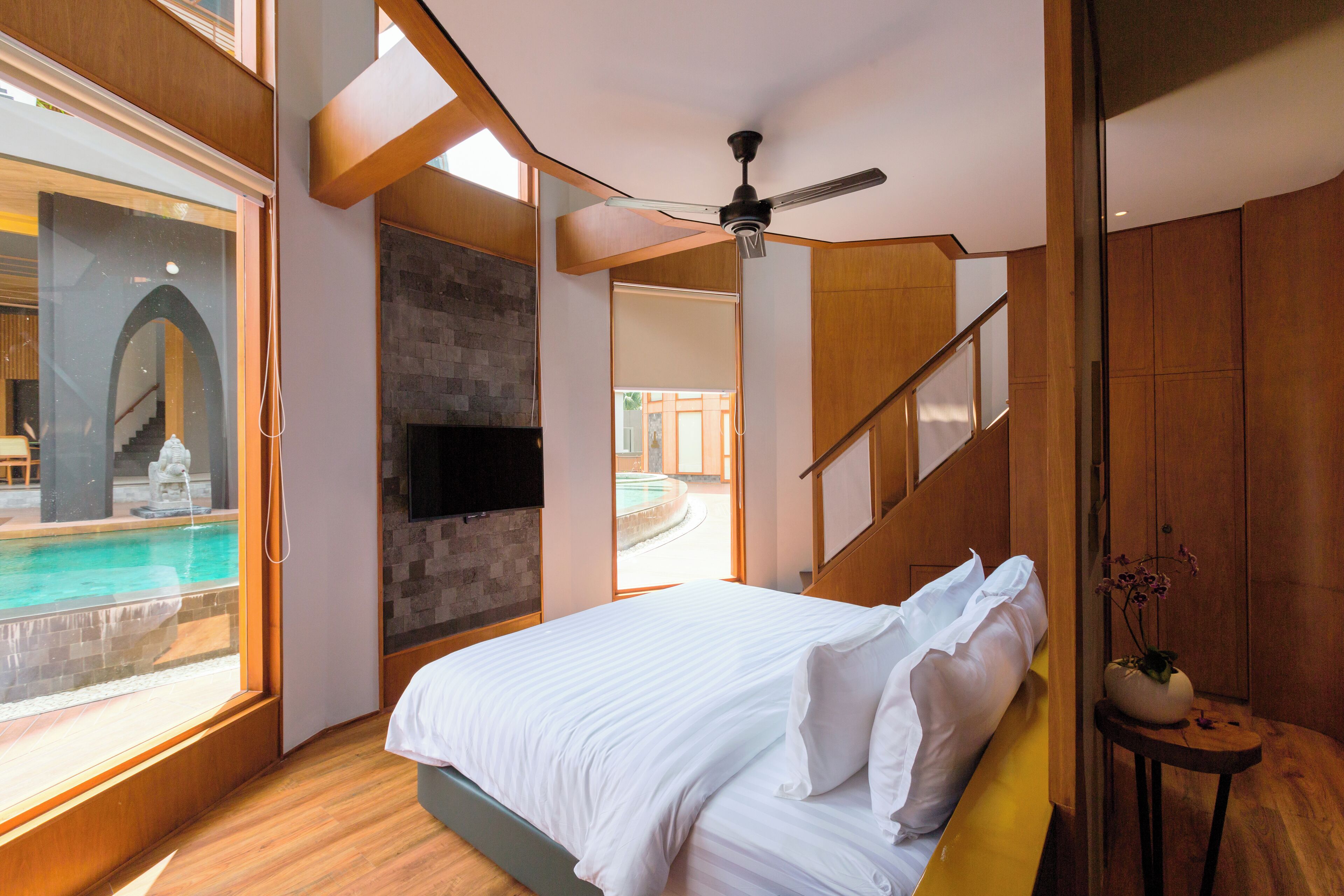 A modern bedroom with a large bed, a ceiling fan, a view of a swimming pool, and a stone fireplace.