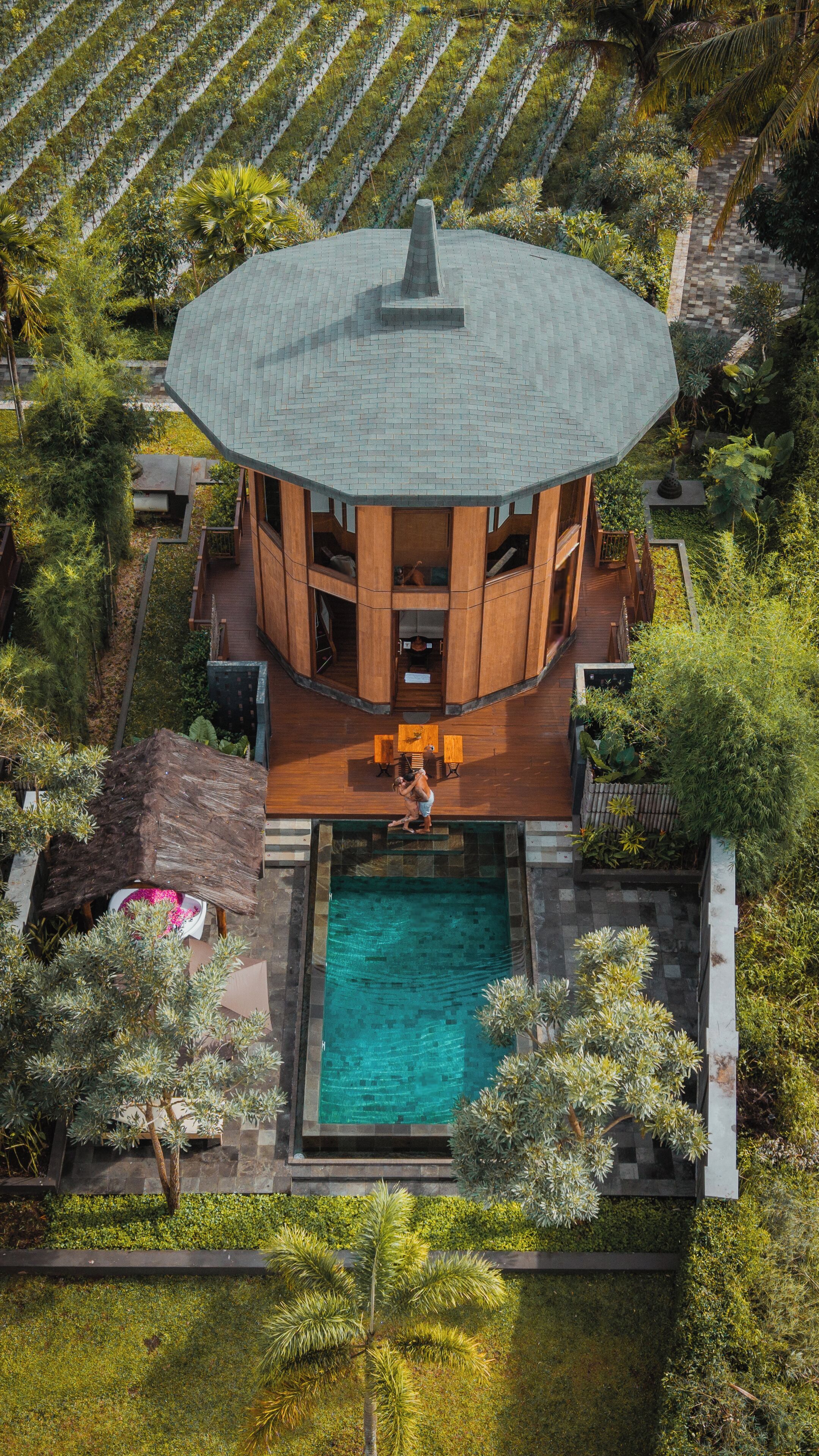A circular building with a central pool, surrounded by lush greenery and a vineyard in the background.