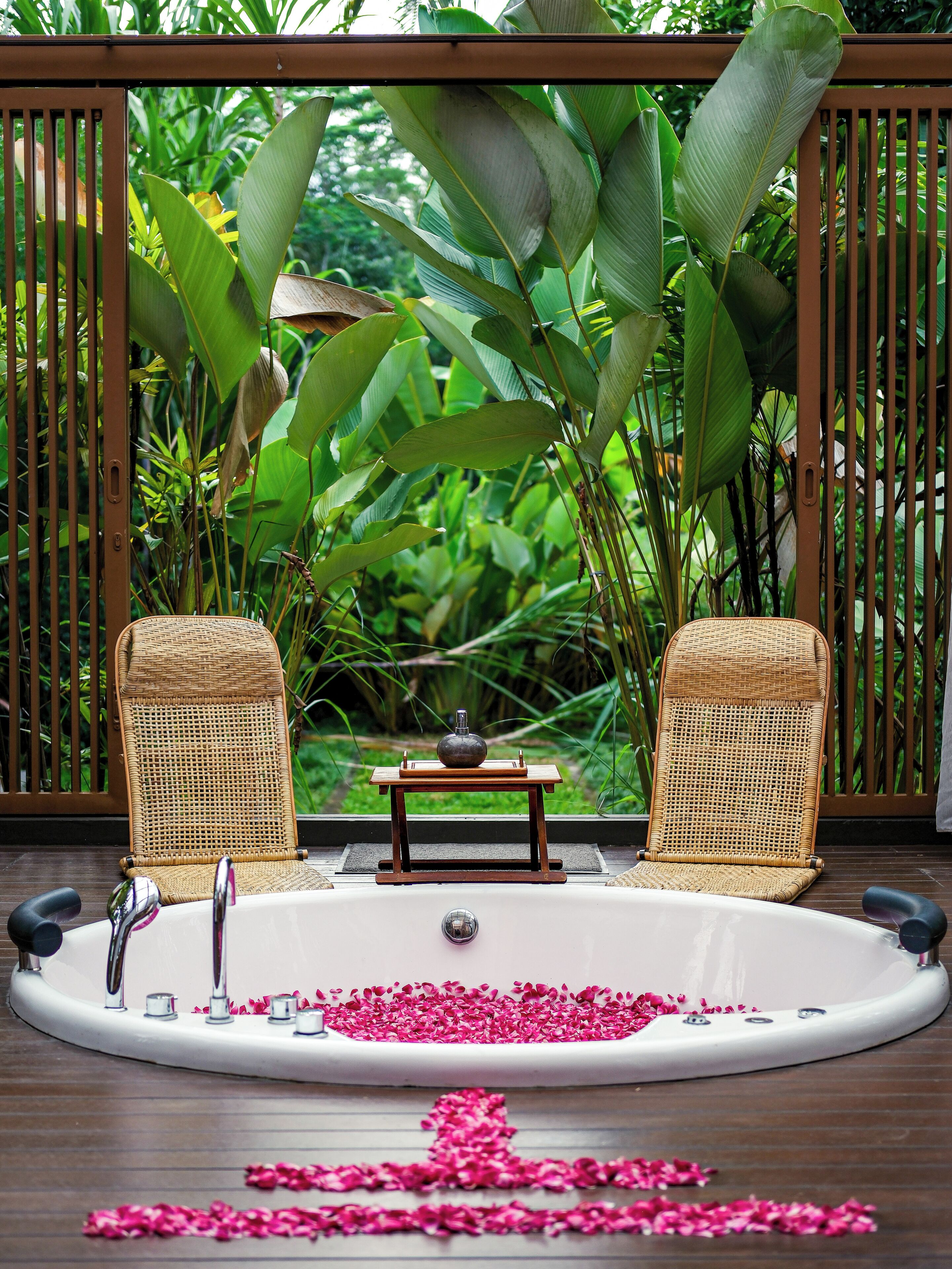A spa area with a bathtub filled with rose petals, two wicker chairs, and a small table with a vase.