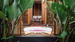 A spa area with a wooden bench, a bathtub filled with rose petals, and lush green plants surrounding the space. - Hotel Le Temple (Borobudur)