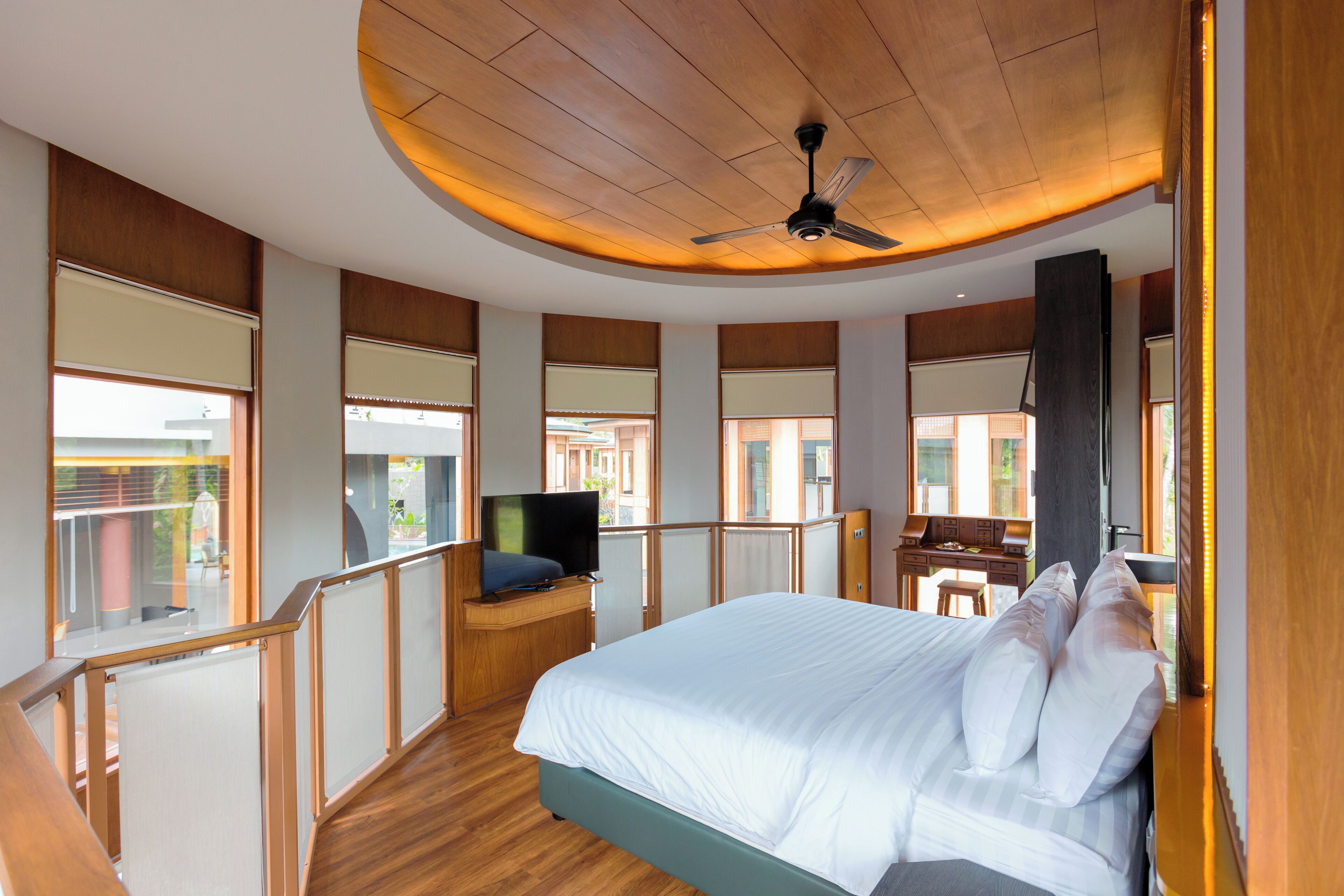 A modern hotel room with a large bed, a desk with a computer, a TV, and a balcony with a view.