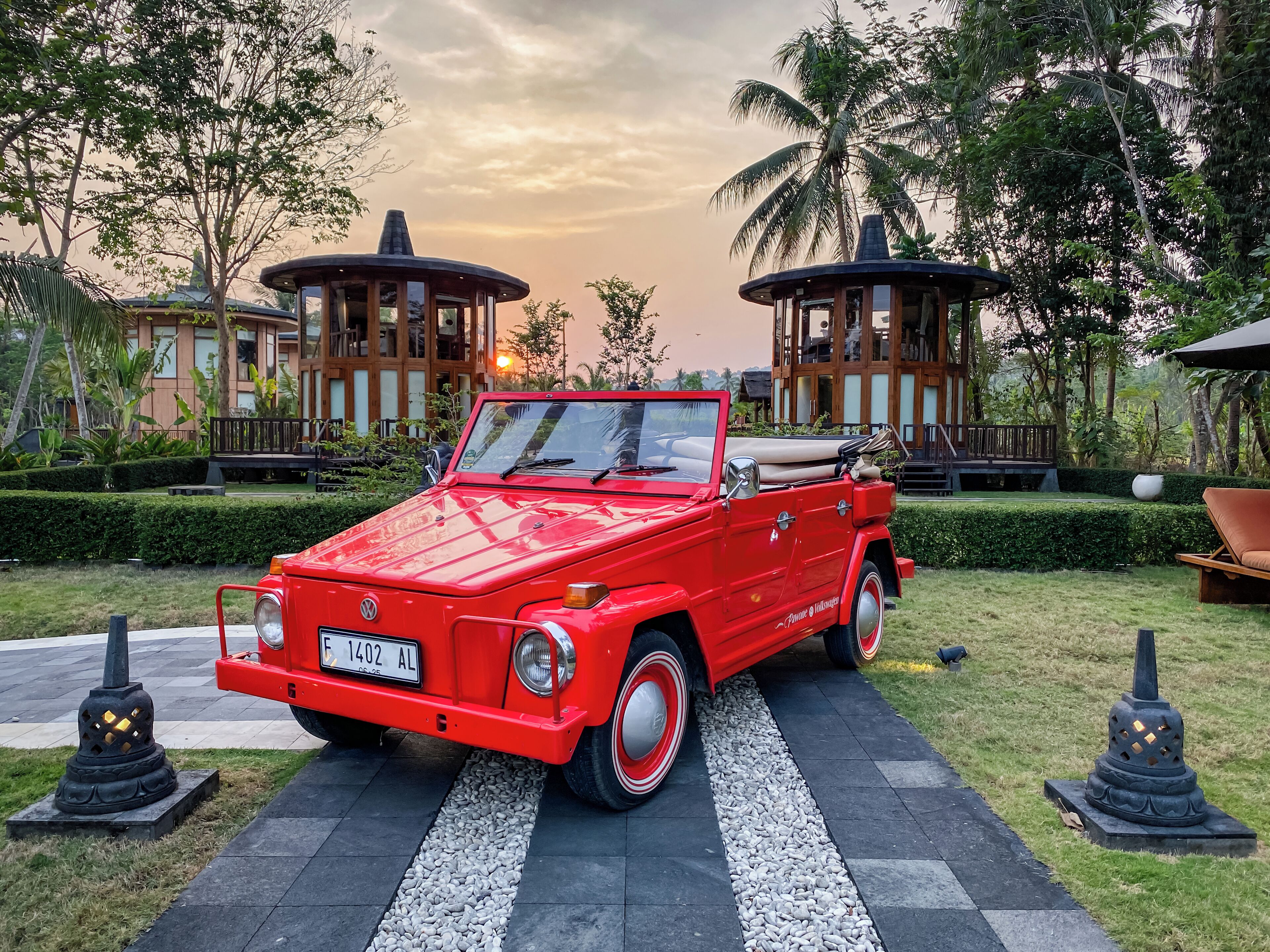 A red vintage car parked on a paved path in a garden setting with two wooden gazebos and lush greenery.