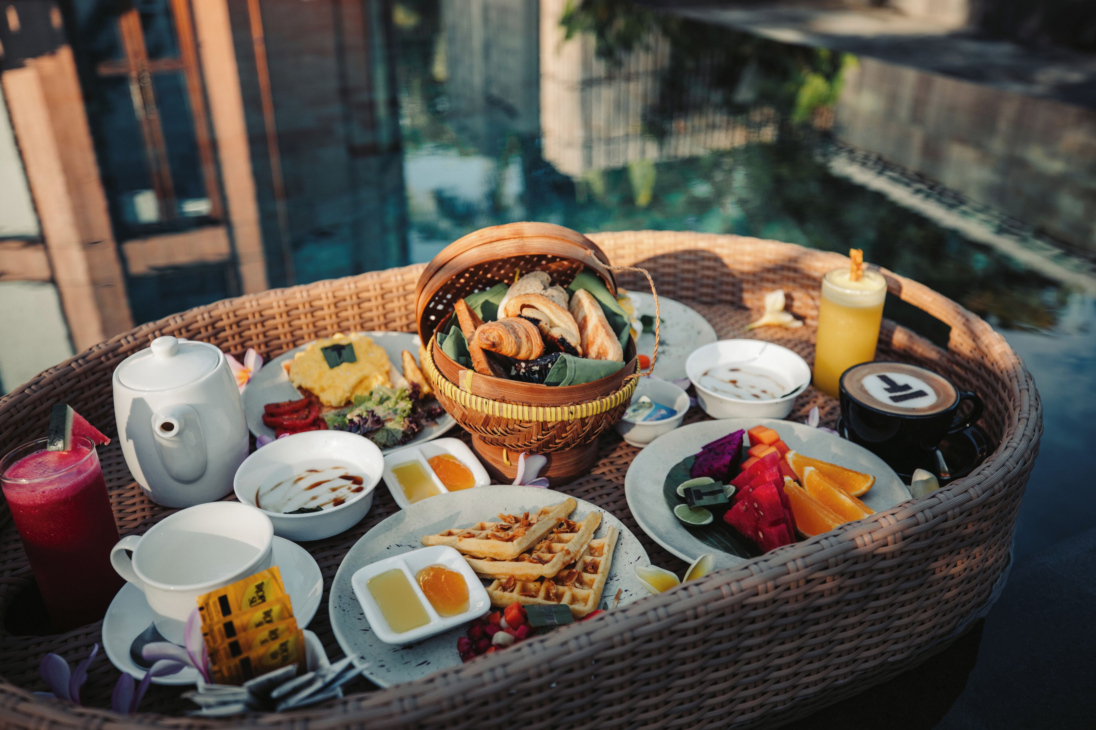En bricka av rotting med ett urval av frukosträtter, inklusive våfflor, bakverk och frukt, placerad nära en pool.
