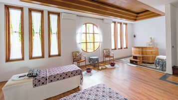 A spacious room with wooden floors, large windows, a wooden bathtub, and two beds with patterned bedding.