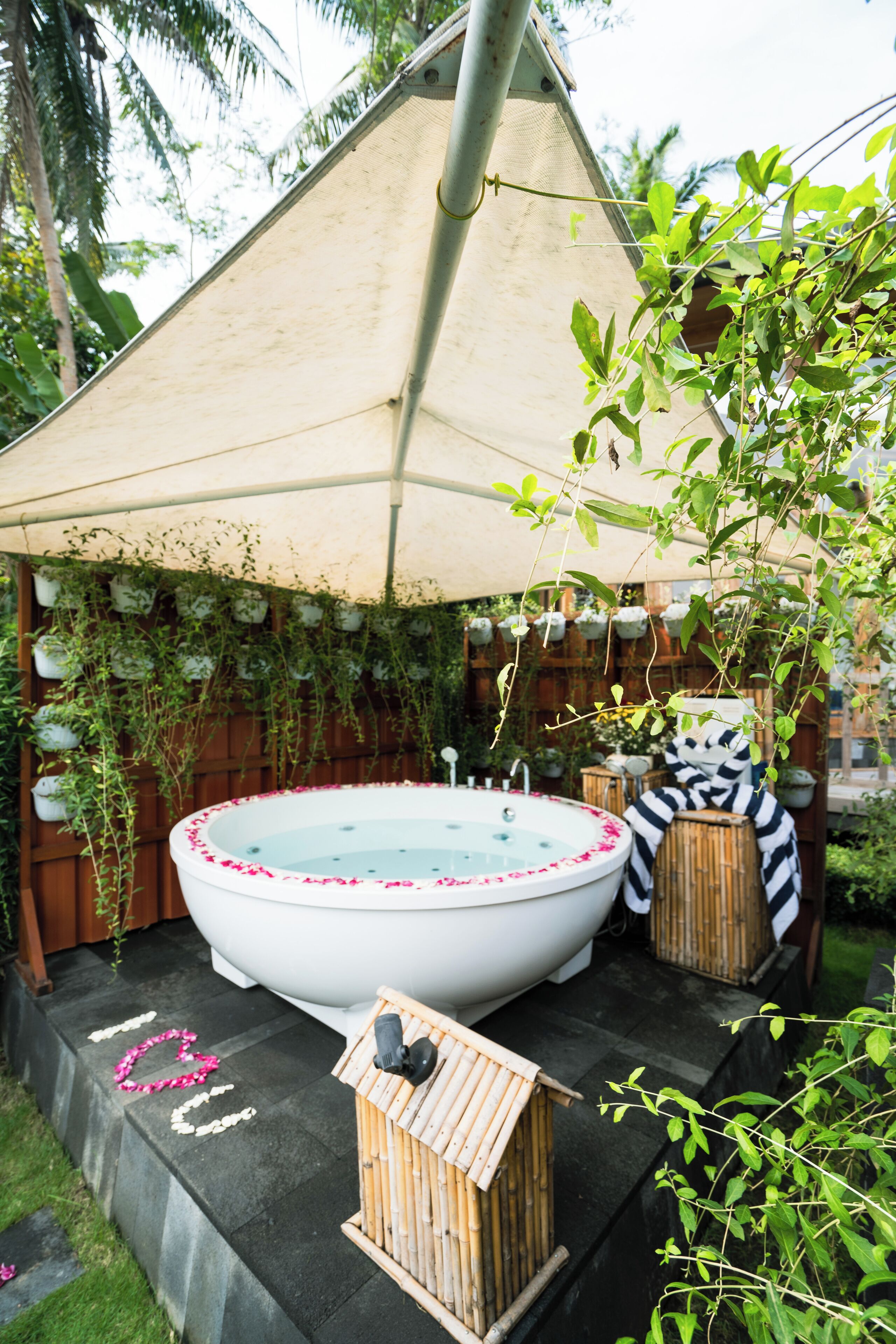 Een hottub met een witte buitenkant en roze bloemblaadjes rond de rand, omgeven door weelderige begroeiing en een houten omheining.