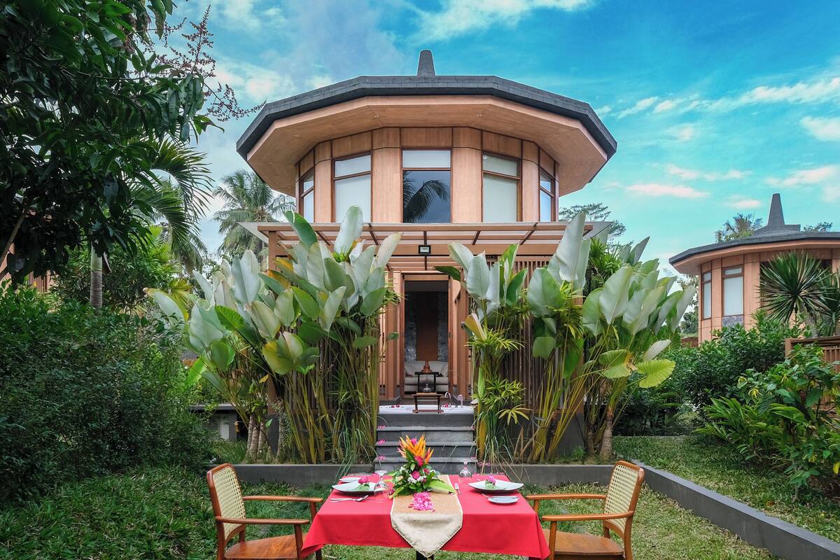 Comedor con una mesa roja dispuesta para cuatro, rodeado de abundante vegetación y un edificio grande al fondo.