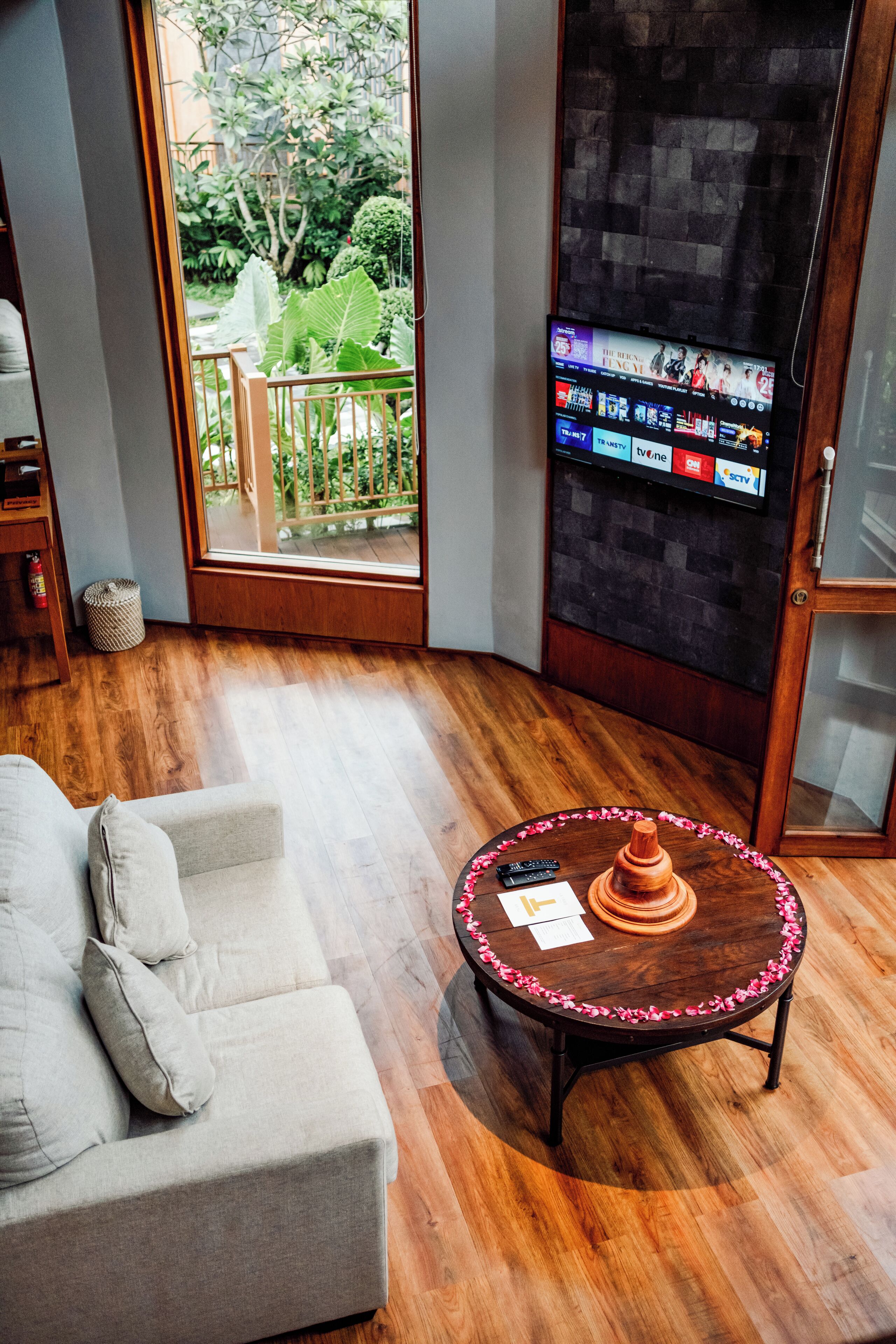 Una sala de estar moderna con piso de madera, un sofá gris, una mesa redonda con un control remoto y papeles, y un televisor mostrando varios canales.