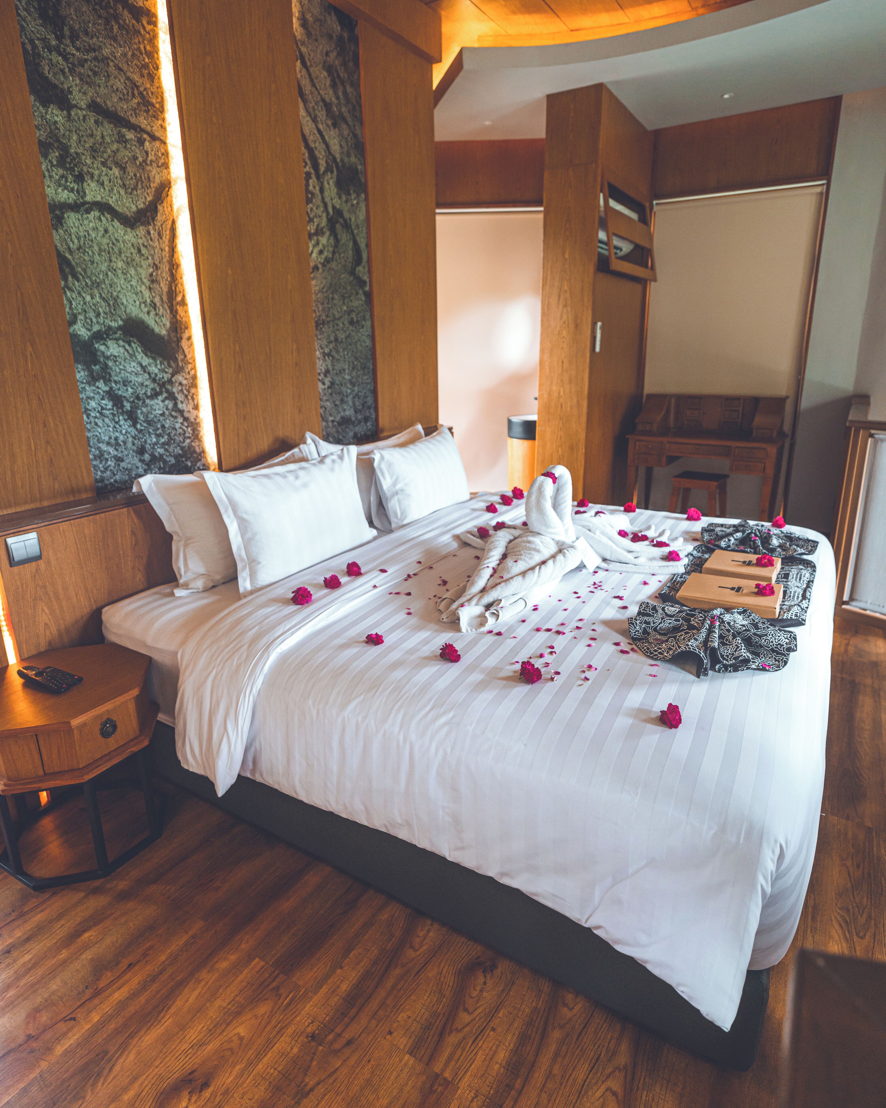 A neatly made bed with white linens and rose petals, a wooden nightstand with a remote control, and a wooden floor.