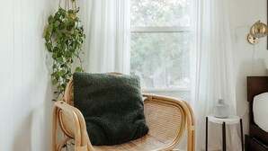 A room with a wicker chair, a patterned rug, a bedside table with a lamp, and a window with curtains. - The Haven • Outdoor Tub • Backyard Oasis • As seen on Magnolia Network (Kill Devil Hills)