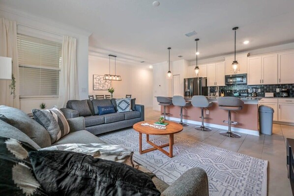 A modern living room with a sofa, coffee table, and bar stools. A kitchen with white cabinets and a black countertop is visible in the background.