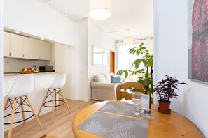 Een moderne keuken met witte kastjes, een eethoek met een ronde tafel, en een woonkamer met een bank en een groot raam.