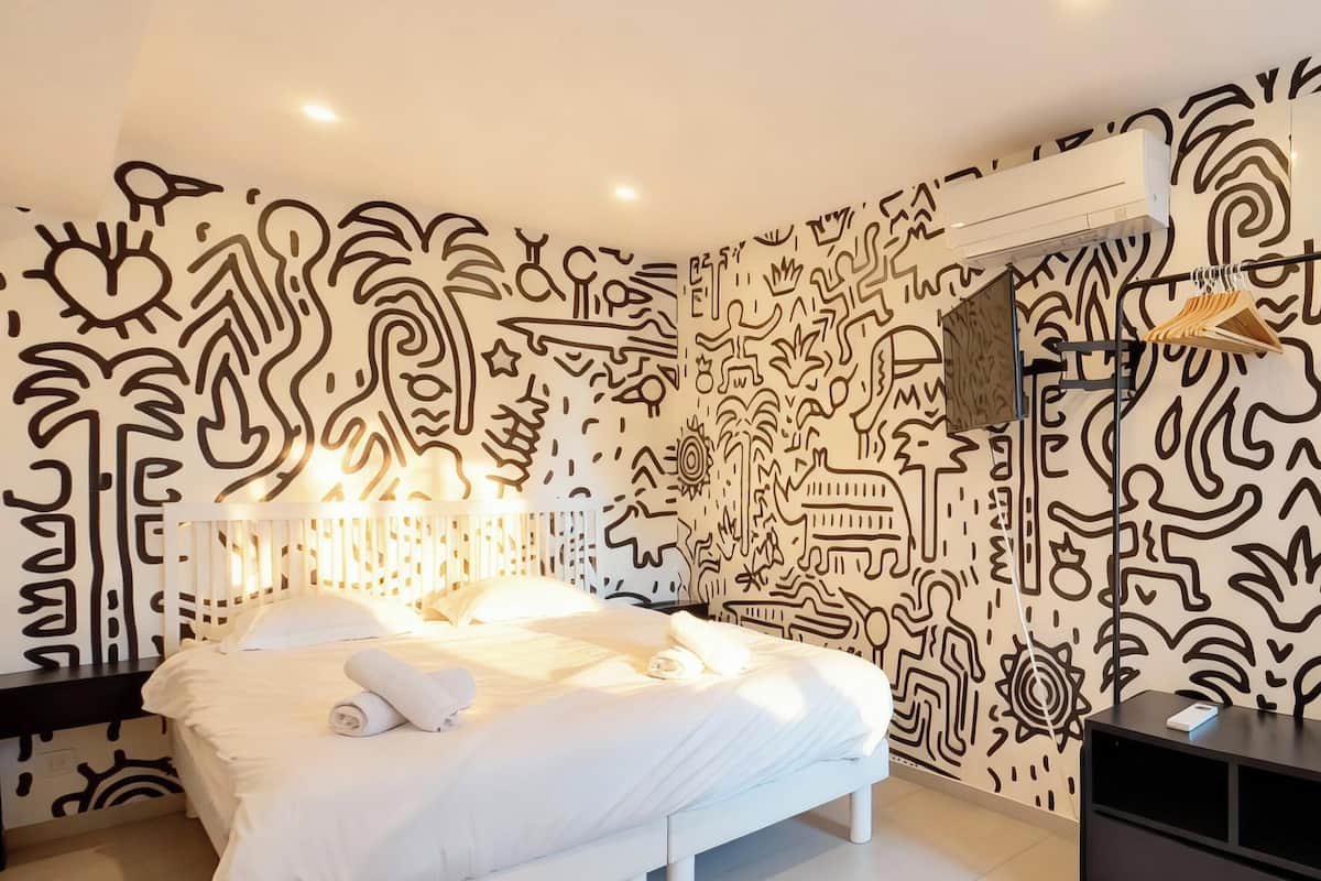 A modern bedroom with a white bed, a nightstand, a dresser, and a wall with abstract black patterns.