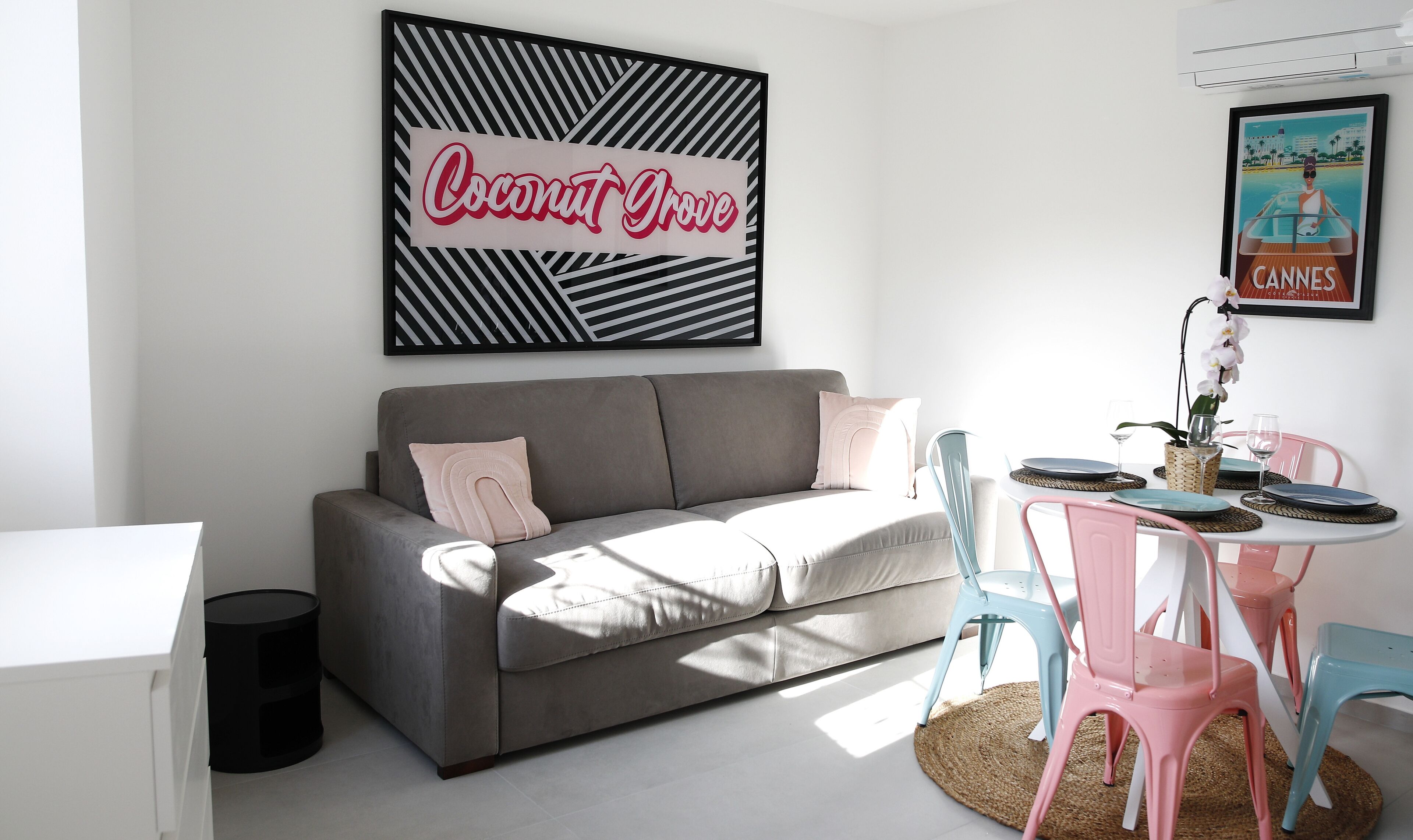 Een woonkamer met een grijze bank, een kleine ronde tafel met stoelen, en wanddecoratie waaronder een bordje met de tekst 'Coconut Grove' en een poster van 'Cannes'.
