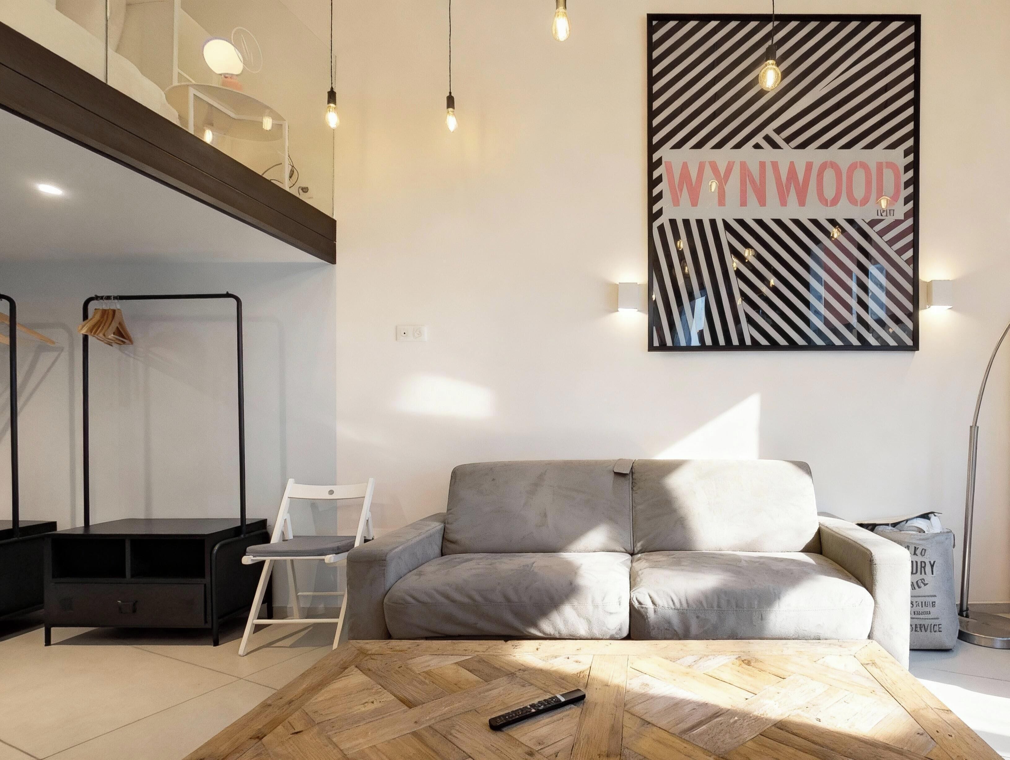 Un salon moderne avec un canapé gris, une table basse en bois et un meuble TV noir. Une œuvre d’art encadrée portant l’inscription « WYNWOOD » est accrochée au mur.