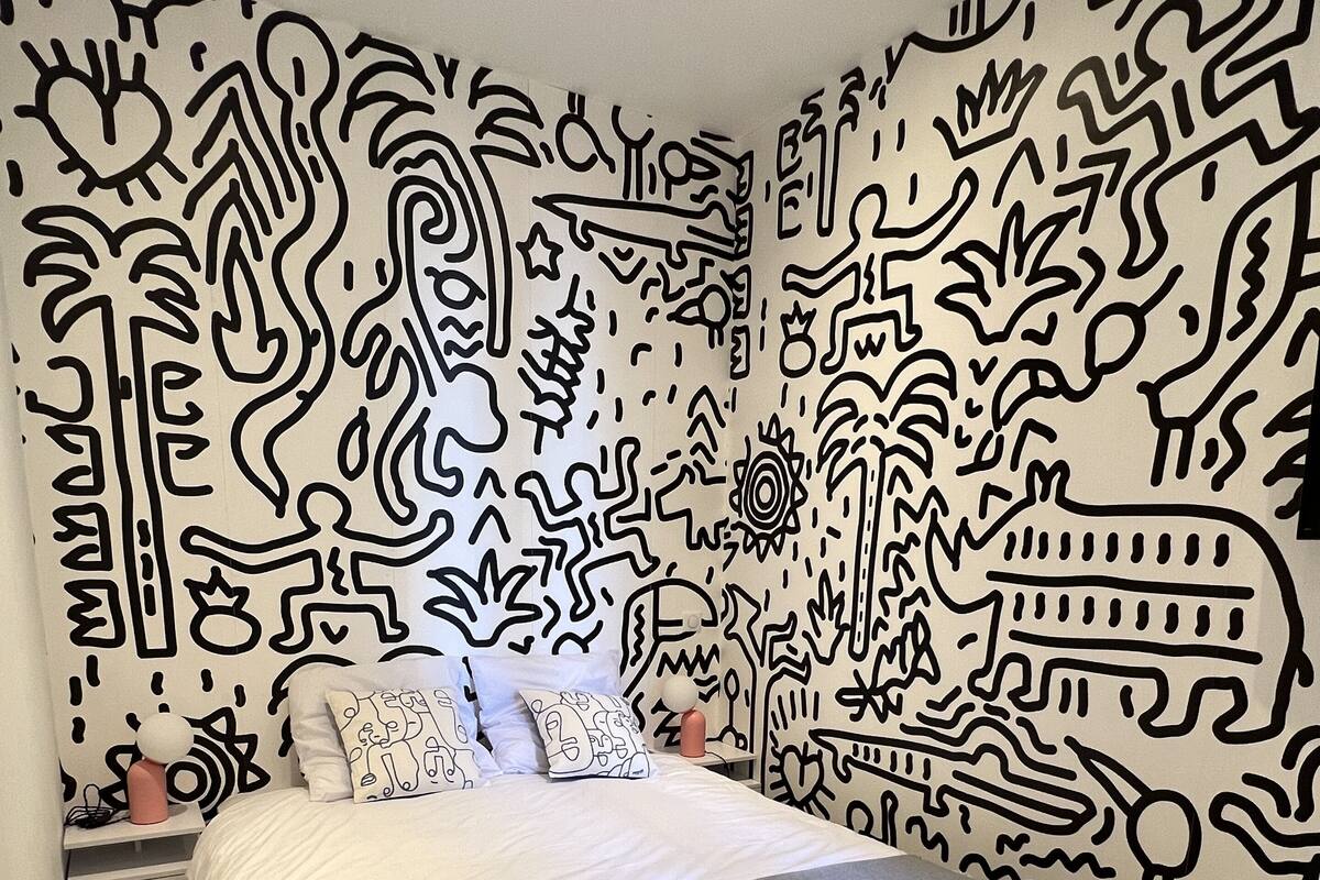 A bedroom with a bed, pillows, a bedside table, and a wall with abstract black patterns.