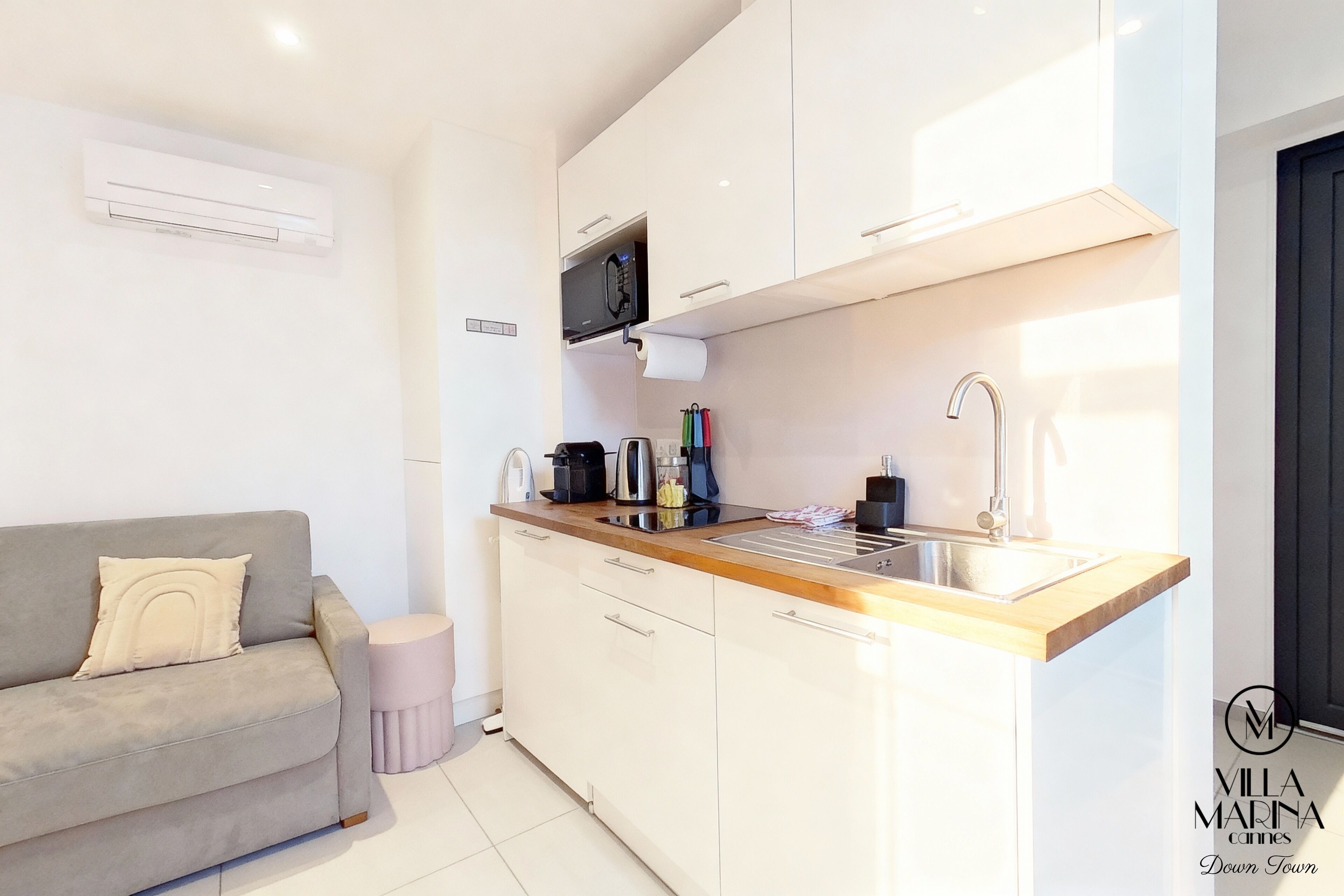 A modern kitchen with white cabinets, a grey sofa, and a small round stool.
