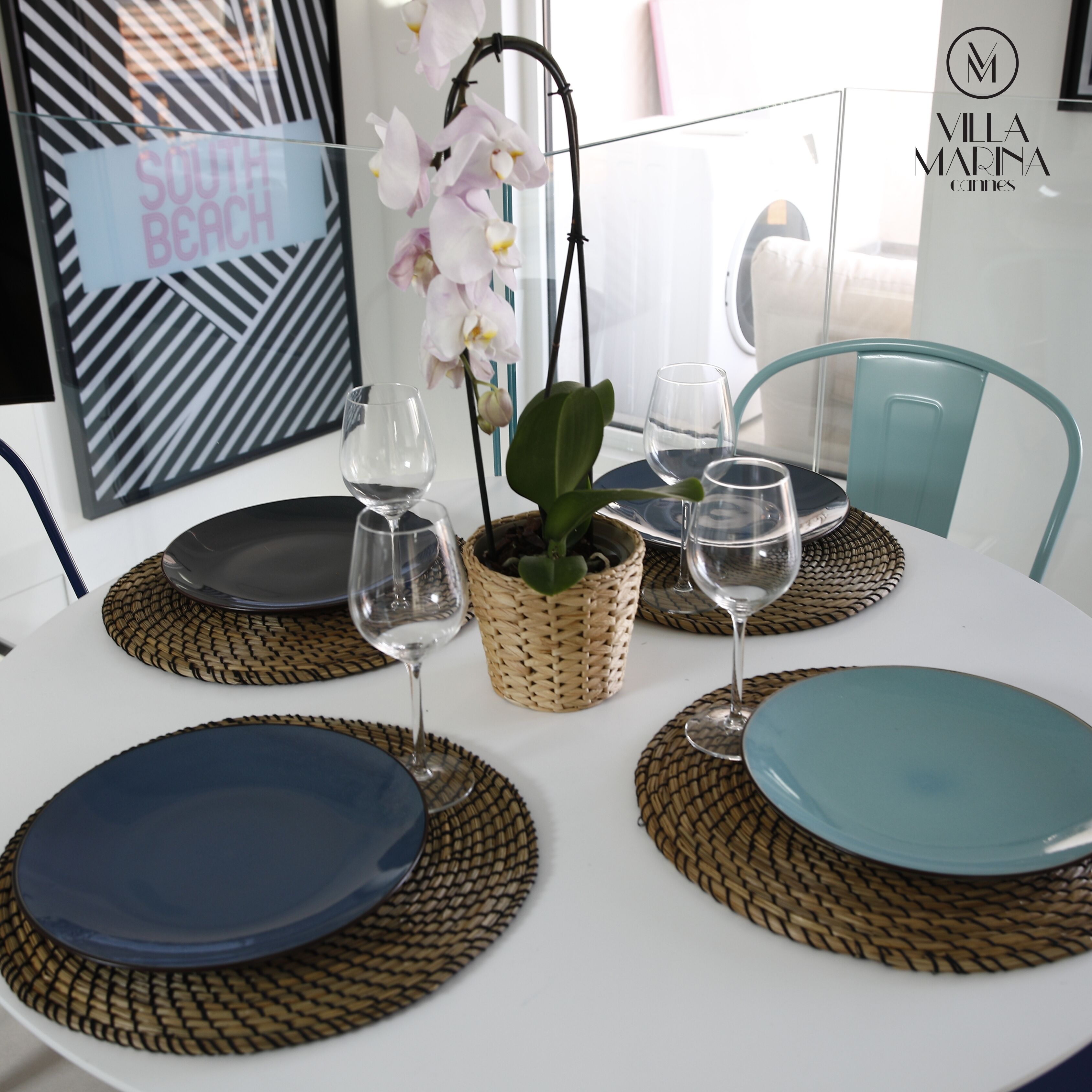 Mesa para comer puesta con platos, vasos y una orquídea en maceta.