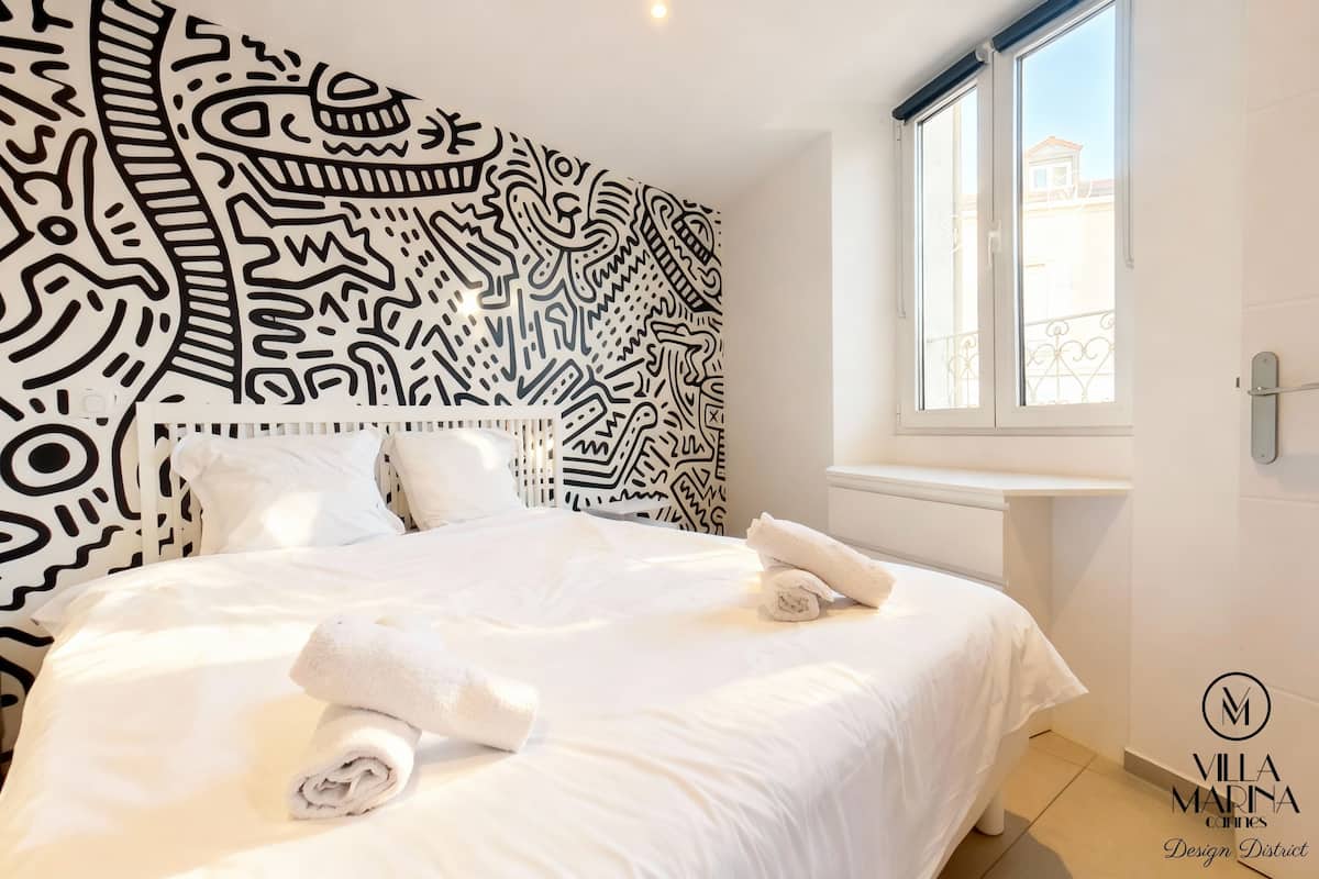 A hotel room with a large bed, white bedding, a patterned headboard, and a window with a view of buildings.