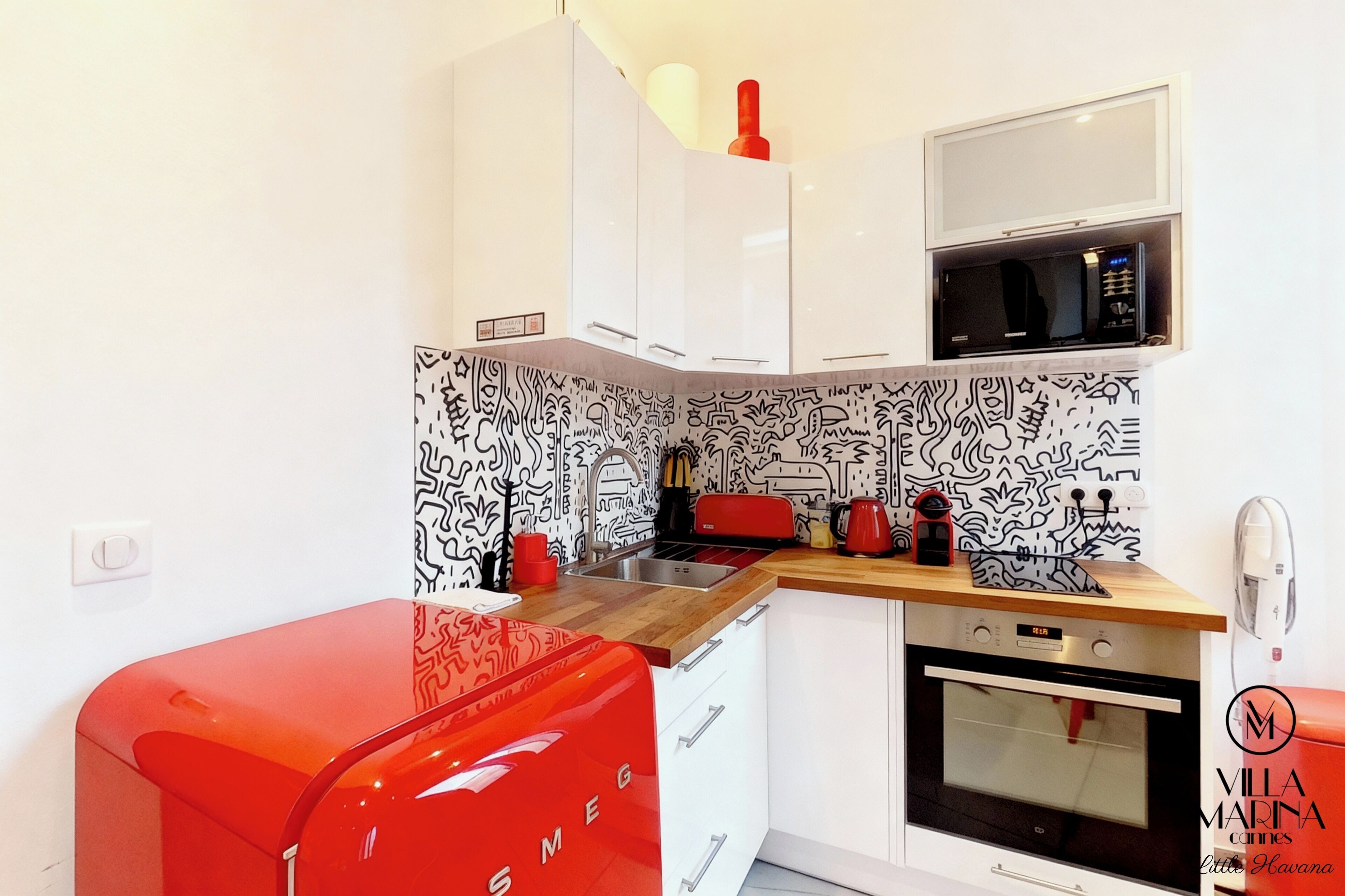 Une cuisine compacte avec des meubles blancs, un four à micro-ondes, un appareil électroménager rouge et un pare-feu décoratif.